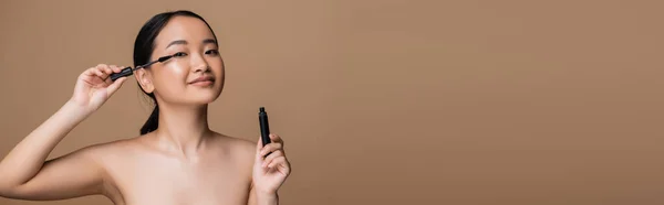 Pretty asian woman with naked shoulders holding mascara isolated on brown, banner — Stockfoto