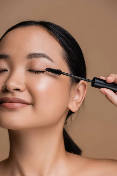 Cropped view of young asian woman holding applicator for mascara isolated on brown - foto de stock
