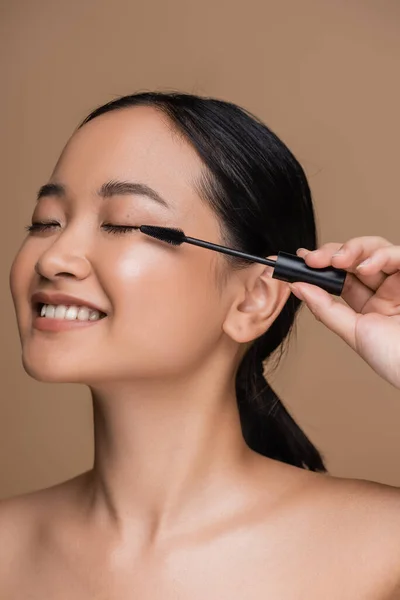 Cheerful asian woman with naked shoulders holding mascara applicator isolated on brown — Stockfoto