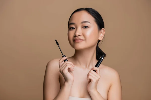 Pretty asian woman holding mascara and looking at camera isolated on brown — Stock Photo