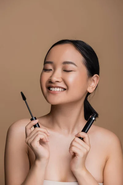 Cheerful asian woman with naked shoulders holding mascara isolated on brown — Stockfoto