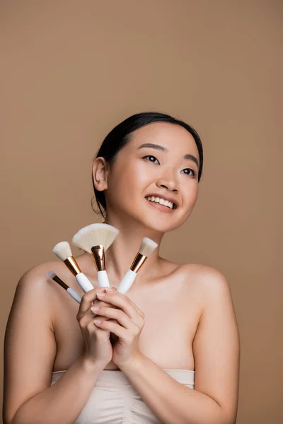 Positive asian model in top holding cosmetic brushes isolated on brown — Stockfoto