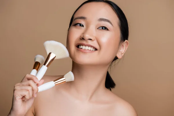 Pretty asian woman holding cosmetic brushes and looking away isolated on brown — Stock Photo