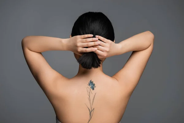 Back view of tattooed woman with naked shoulders touching ponytail isolated on grey — Foto stock
