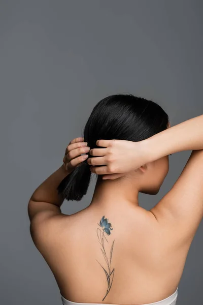 Вид збоку молодої азіатки з татуюванням на спині торкається волосся ізольовано на сірому — стокове фото