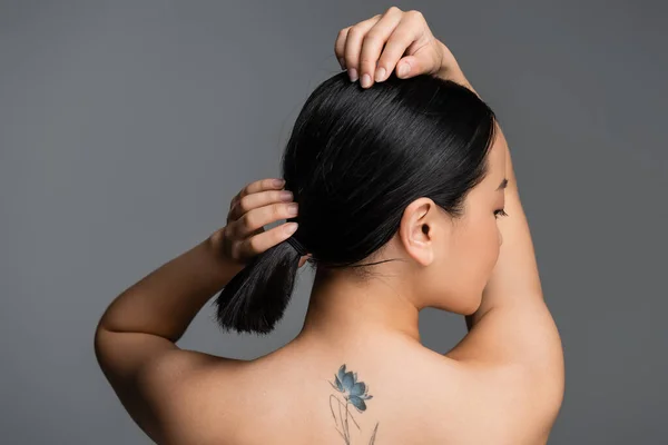 Side view of pretty asian woman with tattoo on back touching hair isolated on grey - foto de stock