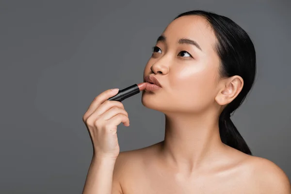 Pretty asian woman applying lipstick and looking away isolated on grey — Stockfoto