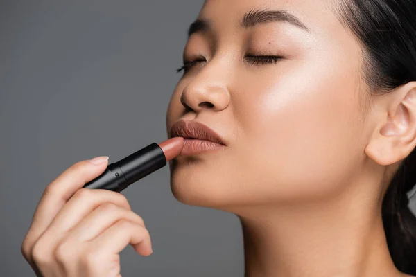 Young asian woman with makeup applying lipstick isolated on grey — Photo de stock