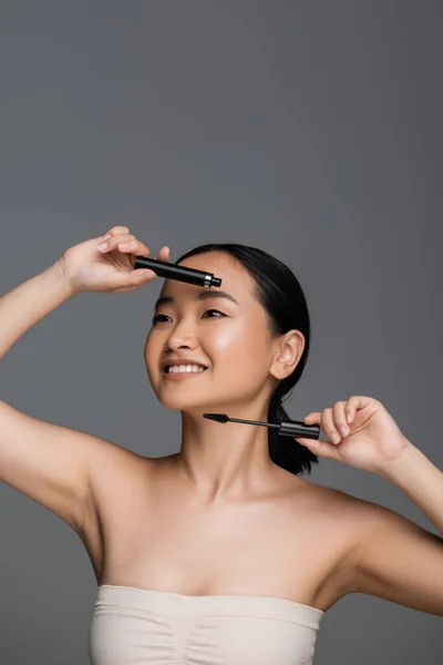 Cheerful asian woman in top holding mascara and eyelash brush isolated on grey - foto de stock