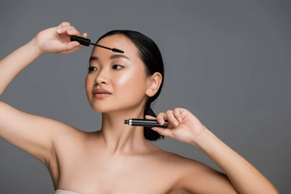 Brunette asian woman with naked shoulders and perfect skin holding black mascara isolated on grey — Stockfoto