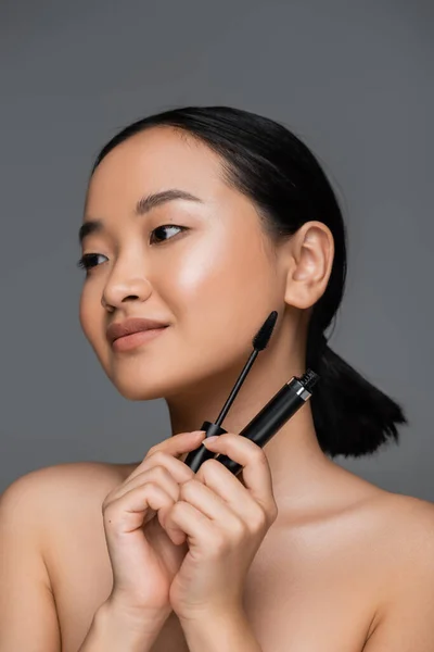 Pleased asian woman with natural makeup holding mascara isolated on grey - foto de stock