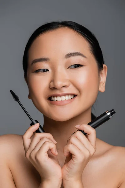 Happy asian woman with bare shoulders and natural makeup holding mascara isolated on grey - foto de stock