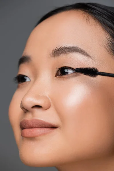 Close up view of asian woman with makeup foundation applying black mascara isolated on grey — Stockfoto