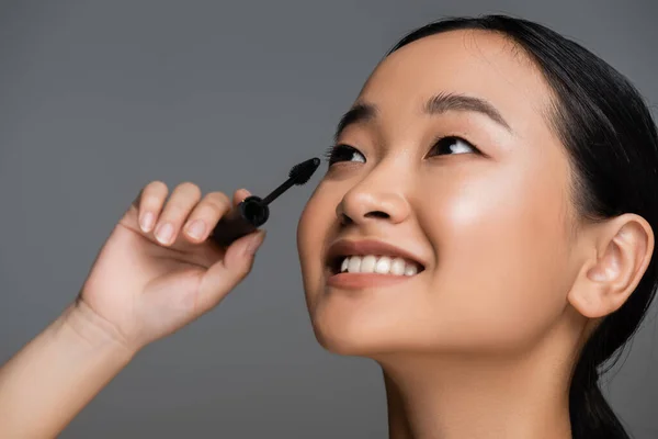 Portrait of young asian woman with perfect smile applying black mascara isolated on grey — Stockfoto