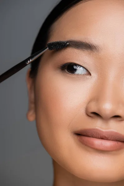 Close up view of cropped asian woman with makeup foundation on face brushing eyebrow isolated on grey — Stockfoto