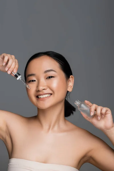 Young asian woman holding bottle and dropper of cosmetic serum while smiling at camera isolated on grey — Stock Photo
