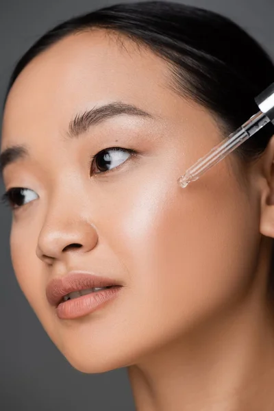 Close up view of asian woman applying moisturizing cosmetic serum during facial pampering isolated on grey — Stock Photo