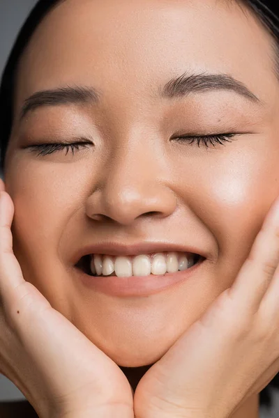 Close up portrait of smiling asian woman with natural makeup touching face while smiling with closed eyes — Stock Photo