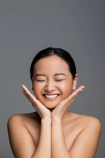 Excited asian woman with closed eyes and nude makeup holding hands near face isolated on grey — Stock Photo