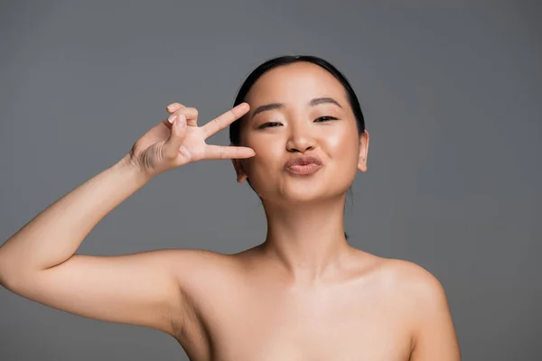 Asian woman with bare shoulders pouting lips and showing victory gesture near face isolated on grey — Stock Photo