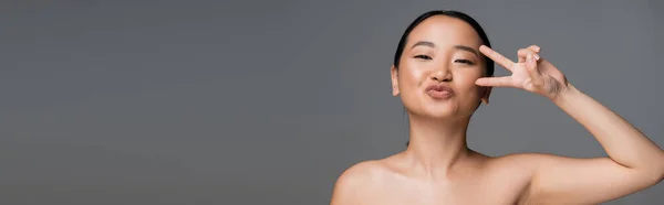 Young asian woman with bare shoulders showing victory sign and pouting lips isolated on grey, banner — Stockfoto