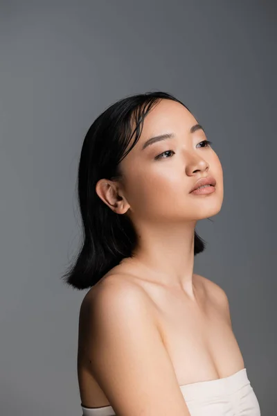 Pretty asian woman with bare shoulders and nude makeup looking away isolated on grey — Stockfoto