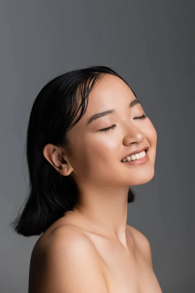Young asian woman with bare shoulders and nude makeup smiling with closed eyes isolated on grey - foto de stock