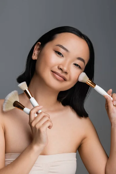 Усміхнена азіатська жінка тримає косметичні щітки і наносить порошок для обличчя ізольовано на сірий — стокове фото