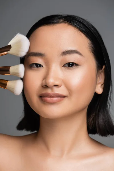Portrait of brunette asian woman with natural makeup looking at camera near different cosmetic brushes isolated on grey — Stockfoto