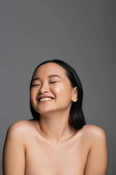 Cheerful asian woman with bare shoulders and natural makeup smiling with closed eyes isolated on grey — Stock Photo