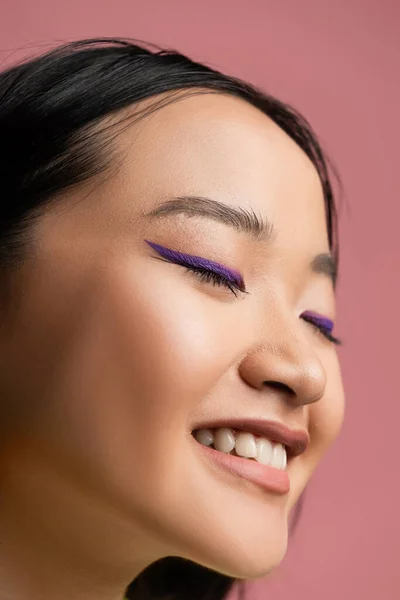Крупним планом молода азіатка з макіяжем і блакитною підводкою посміхаючись із закритими очима ізольовані на рожевому — стокове фото