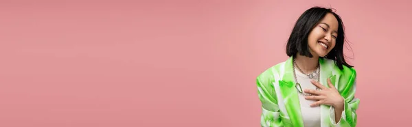 Збуджена азіатська жінка в зелено-білій куртці торкається грудей, сміючись із закритими очима ізольована на рожевому, банер — стокове фото