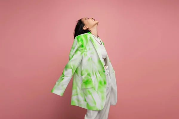 Молода і щаслива азіатська жінка в зеленому і білому кольорі оверсайз Блейзер позує з піднятою головою ізольовано на рожевому — стокове фото