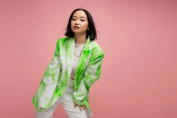 Молода азіатка в модній зелено-білій куртці дивиться на камеру, позує ізольовано на рожевому — стокове фото