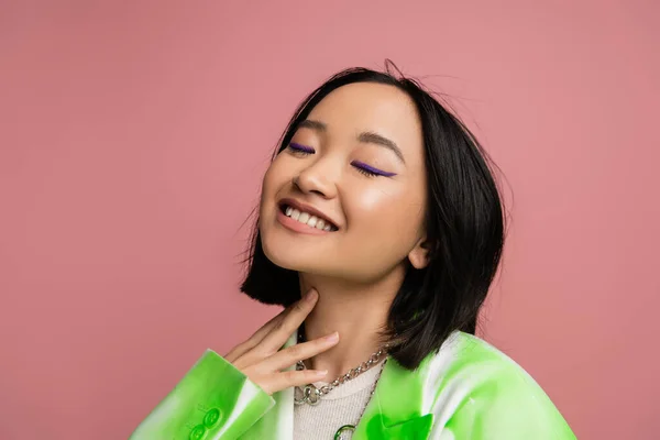Happy asian model in metal necklaces and makeup posing with closed eyes isolated on pink — Stock Photo