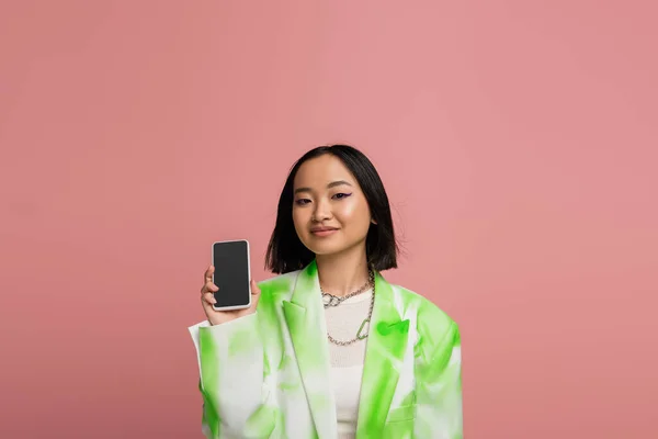 Усміхнена азіатська жінка в модній куртці, що показує смартфон з порожнім екраном ізольовано на рожевому — стокове фото