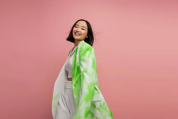 Happy asian woman in trendy oversize blazer smiling at camera while walking isolated on pink — Stock Photo