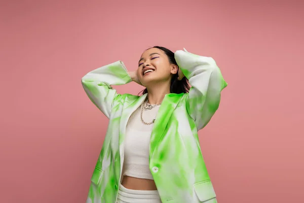 Joyful asian woman in green and white jacket touching hair and posing with closed eyes isolated on pink — Stock Photo