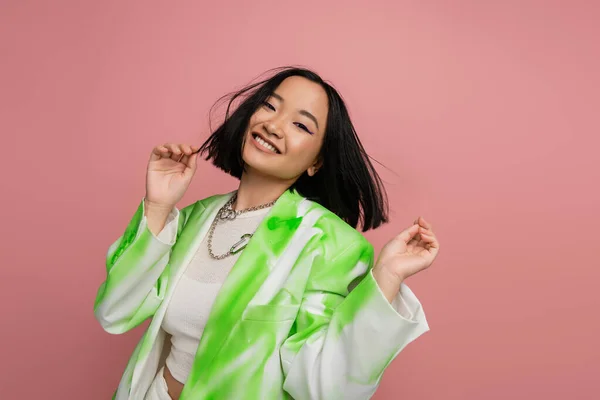 Весела азіатська жінка в стильній зелено-білій куртці і срібних намистах позує ізольовано на рожевому — стокове фото