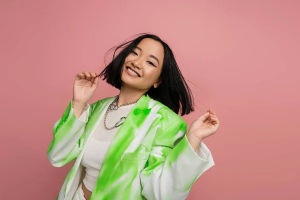 Cheerful asian woman in stylish green and white jacket and silver necklaces posing isolated on pink - foto de stock