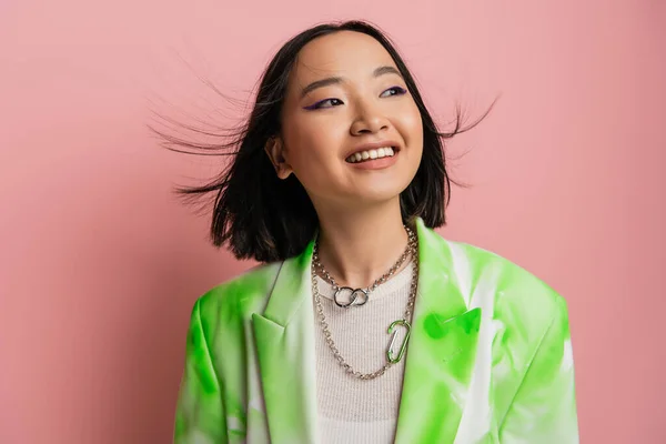 Joyful asian woman in trendy outfit and makeup with blue eyeliner looking away on pink background - foto de stock