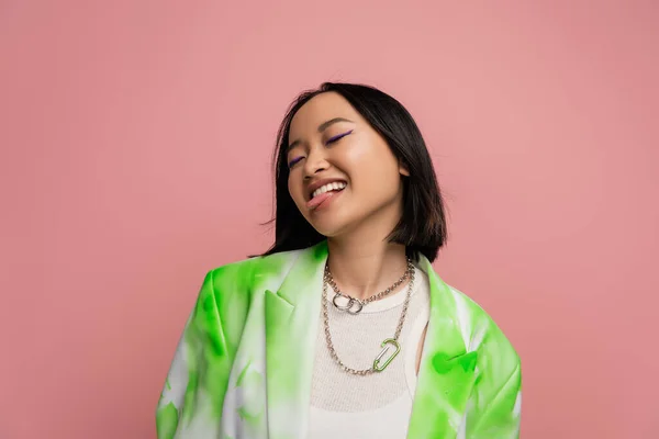 Cheerful asian woman in green and white jacket and metal necklaces sticking out tongue isolated on pink — Stock Photo