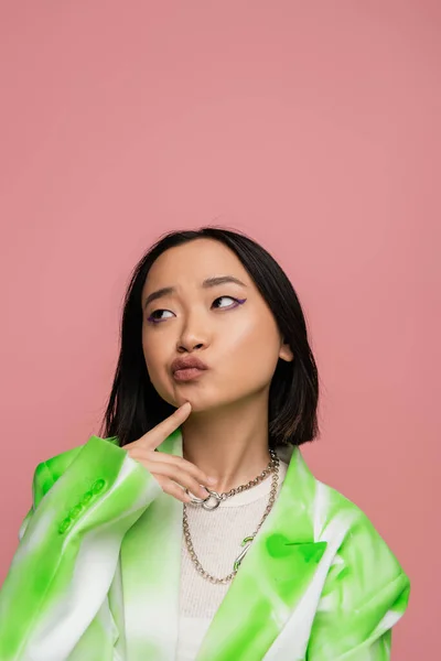Thoughtful asian woman in trendy blazer and metal necklaces touching chin and looking away isolated on pink — Stockfoto