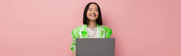 Cheerful asian woman in green and white jacket smiling near laptop isolated on pink, banner - foto de stock