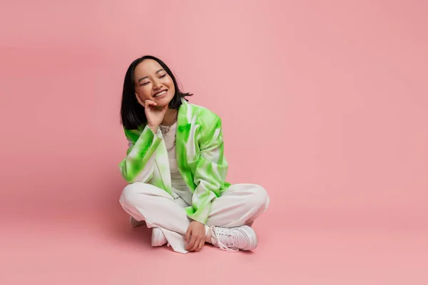 Pleased asian woman in fashionable outfit sitting with crossed legs on pink background — Stock Photo