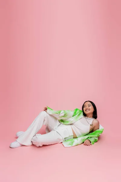 Повна довжина азіатської моделі в білих штанях і модний піджак лежить і дивиться на камеру на рожевому фоні — стокове фото
