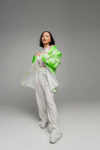 Повна довжина молодої азіатки в штанях і зелено-білій куртці, дивлячись на камеру на сірому фоні — стокове фото