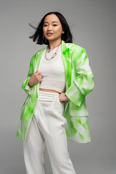 Модна азіатська жінка в зеленому і білому блістері, що стоїть з рукою на стегні ізольована на сірому — стокове фото