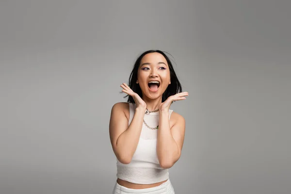 Excited asian woman in white tank top holding hands near face and screaming isolated on grey — Foto stock