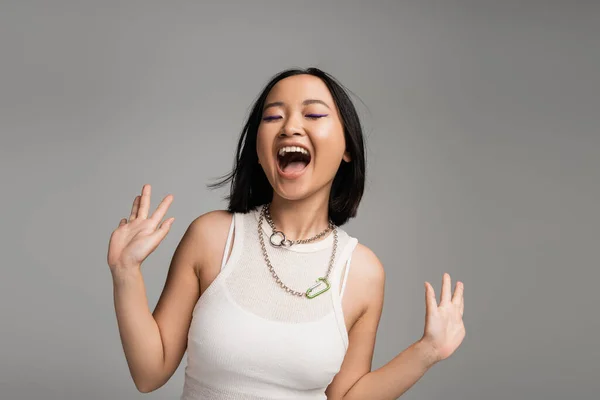 Thrilled asian woman with closed eyes screaming and gesturing isolated on grey — Stockfoto